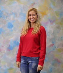 A blonde woman in a red long sleeve shirt and blue jeans stands in front of a colorful photography backdrop.