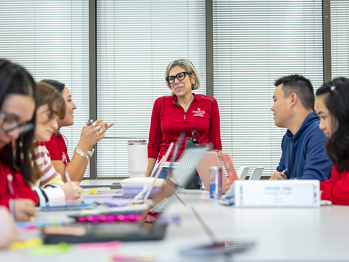 Teacher with glasses and a red shirt standing at a table of college students with laptops