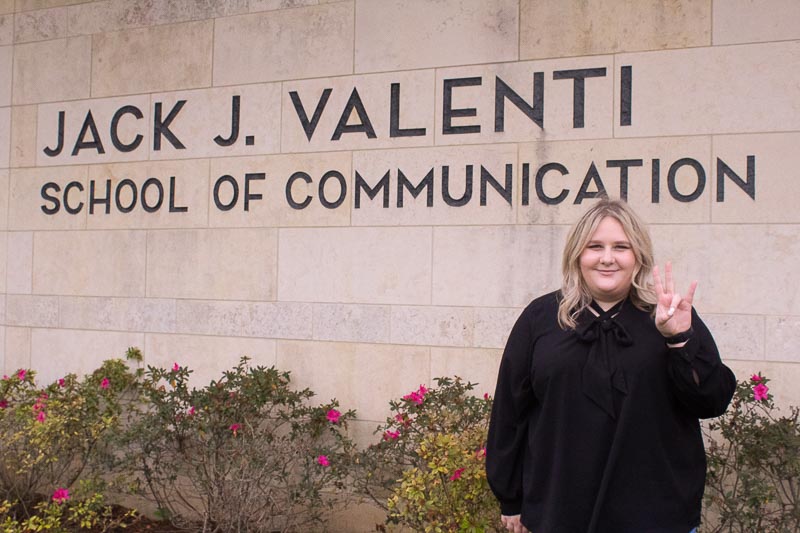 Mandell is standing in front of the Valenti School smiling while throwing up the Coog's sign.