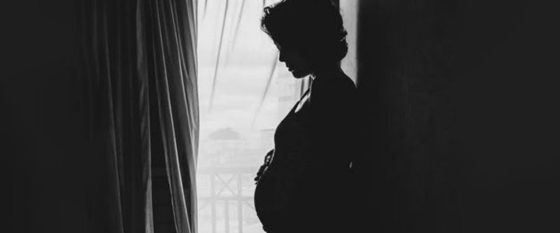 Pregnant and lonely:  an expectant mother looking down at baby bump while standing  in front of a window