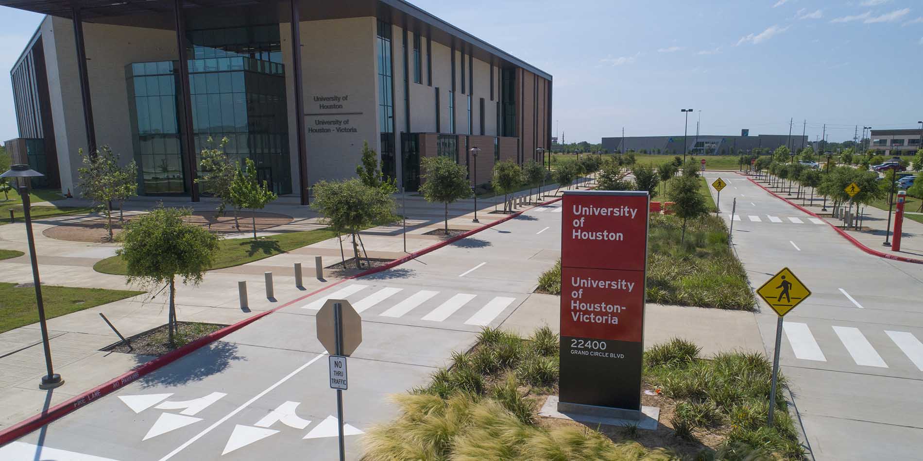 Front entrance of the campus. A red monument sign with "University of Houston" and "University of Houston - Victoria". Further back to the left is a tan brick and glass building.