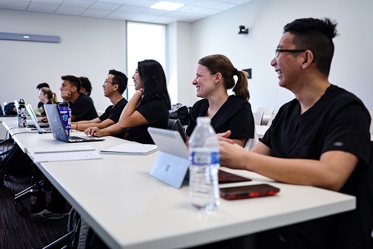 A group of students in black scrubs sit at a row of tables