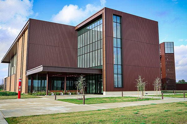 Rear view of a brown three story building. A sidewalk with benches is in the foreground.