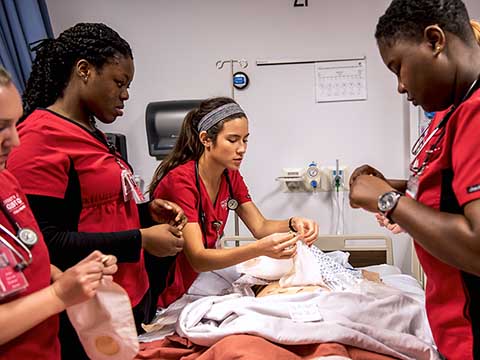 Five nursing students flank a hospital bed while preparing medical supplies.