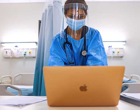 A nurse in a blue surgical gown stands behind an open laptop with her hands on the keyboard.