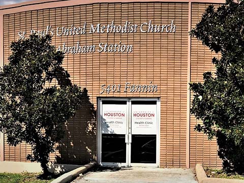 Photograph of the front of a light-brown brick building. The entrance is a set of double glass doors with signs saying "University of Houston College of Nursing Health Clinic" on each.