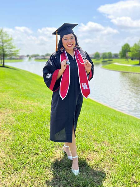 A photo of a hispanic woman wearing graduation regalia on a grass field with a river in the background.