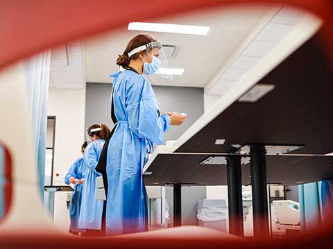 A groupd of nursing students wearing surgical gowns work at tables.