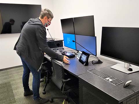 A woman stands over work station with 5 large monitors and flexible gooseneck microphones.