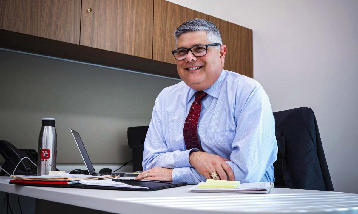 A man in glasses wearing business attire sits at a desk in an office.