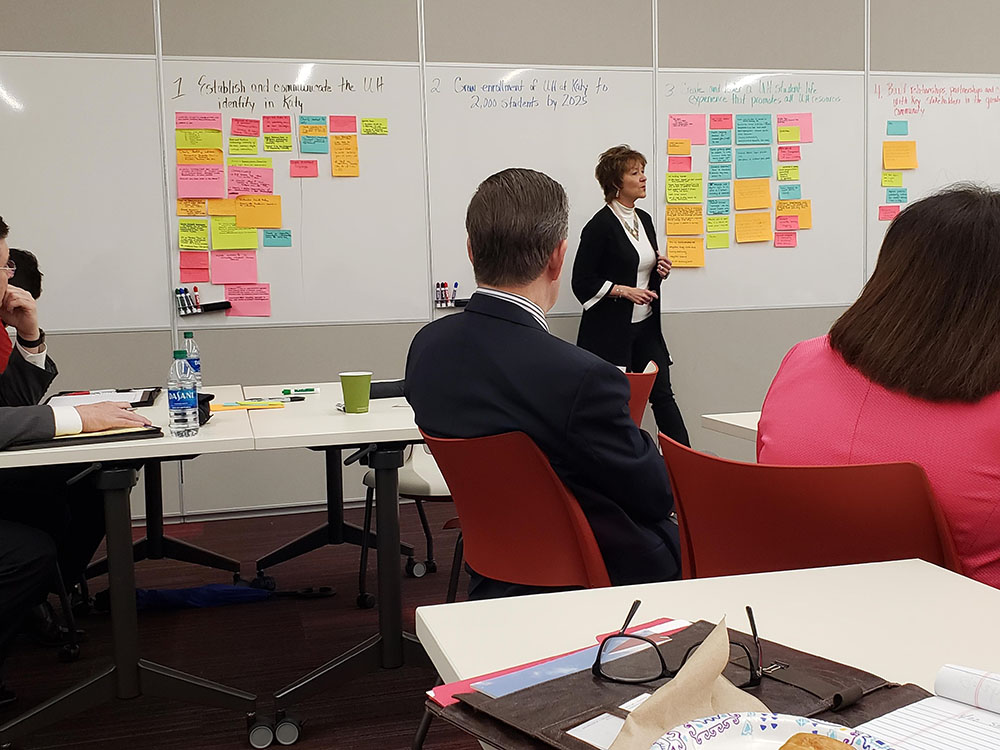 A group of people sitting at tables while a woman facilitates a discussion while standing in front of a white board with hand-written notes