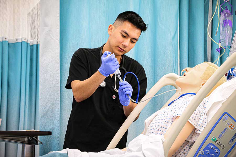 A male nursing student injects a liquid into the intravenous line of a training manikin in a hospital bed.
