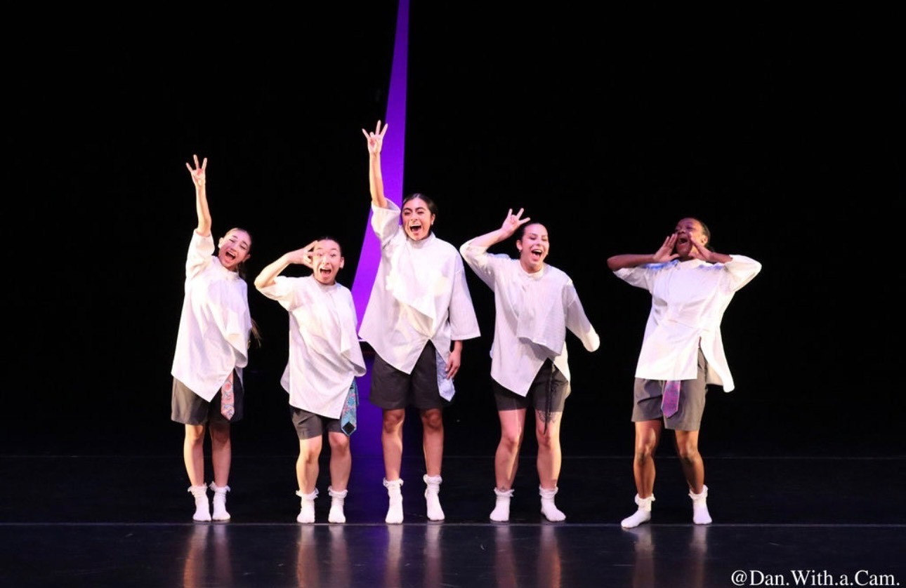 Five performers in white and black costumes stand on a stage.