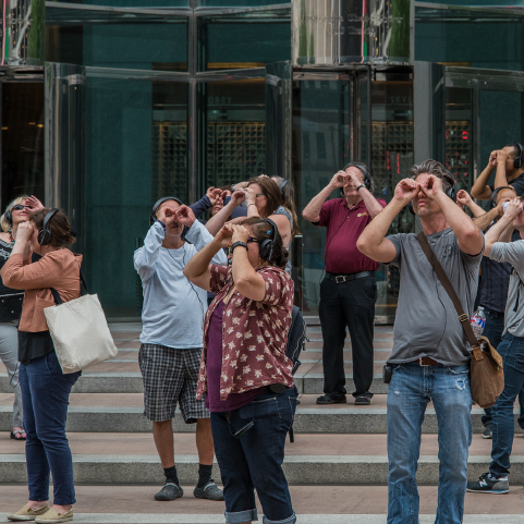 A group of people facing different directions stand on the steps to the entrance to an office building with headphones on, looking through their hands like binoculars.