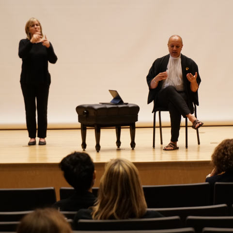 A bald, brown-skinned person seated on a stage wearing a black and white outfit speaks as a blond-haired, white-skinned person wearing all black interprets beside him to on-looking audience members in the foreground.