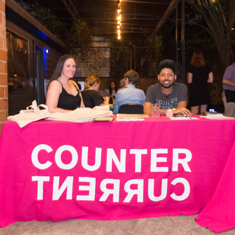 Two people sit behind a table covered with a hot-pink cloth and the words “COUNTER CURRENT” on the front side, situated in a dimly lit public space with people in the background.