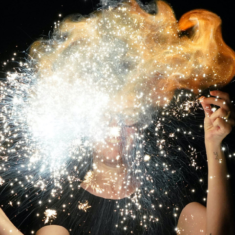 Close-up of a person’s face obscured by an explosion of sparks and fire against a black background.
