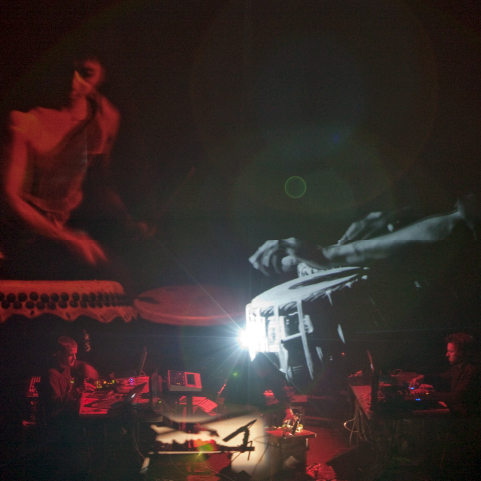 Silhouetted artists on stage use computers and technology to control projections on a large screen above them showing people playing hand drums in red and grey light.