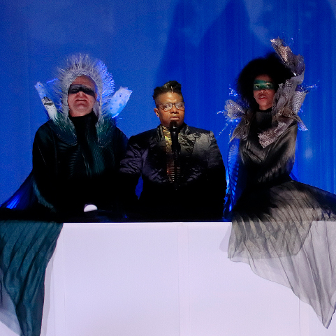 A performance with the main character speaking into a microphone behind a large podium surrounded by people in dark flowing costumes and elaborate headdresses in front of a blue backdrop.