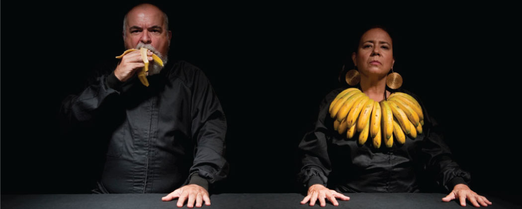Two artists in front of a black backdrop, dressed in black pose behind a black table; the person on the left eats a banana, the person on the right wears a double-layered collar of bananas and large round disk earrings.