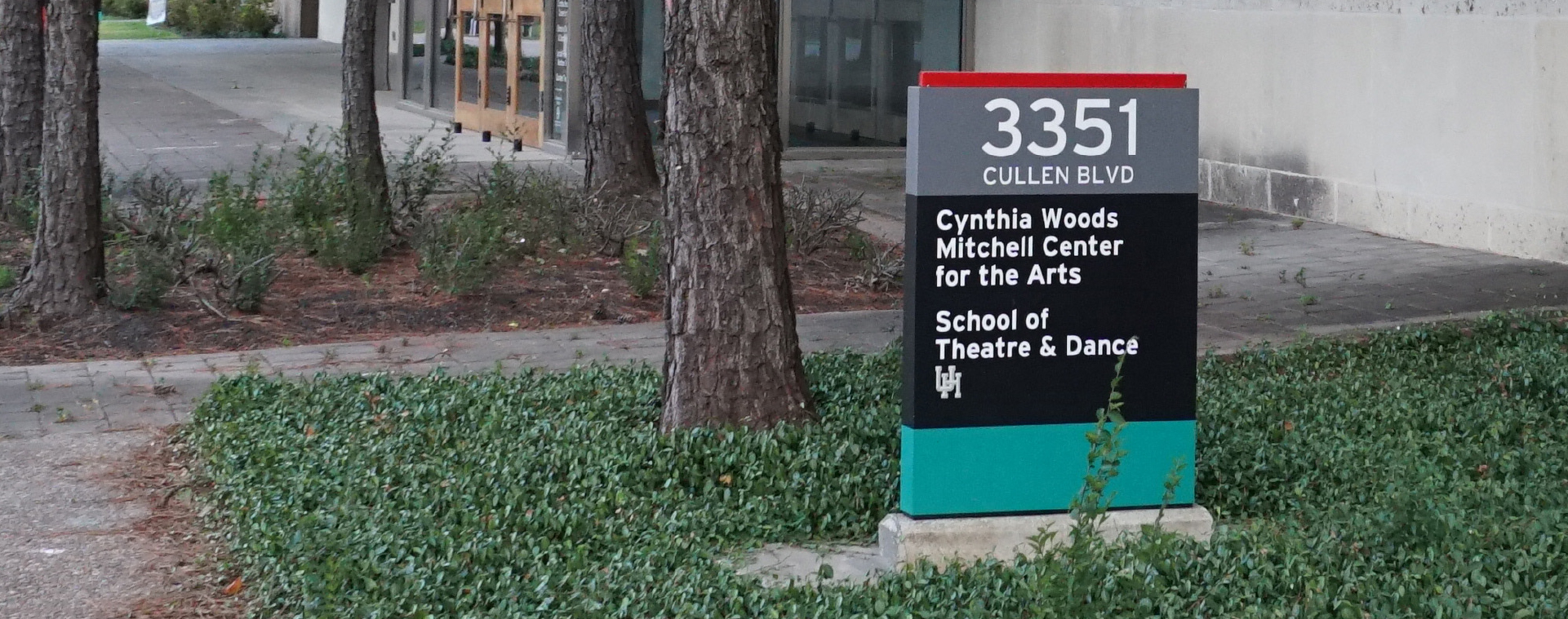 An outdoor sign surrounded by green ground cover reads from top to bottom: “3351 CULLEN BLVD.; Cynthia Woods Mitchell Center for the Arts; School of Theater & Dance; UH.”