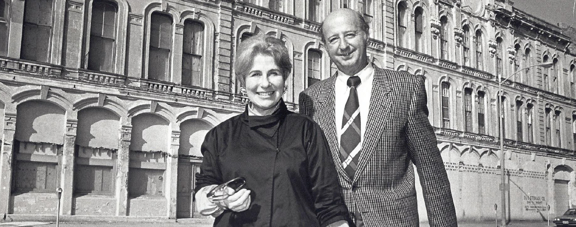 A historic, black and white photo of Cynthia Woods Mitchell and her husband in front of a building in daylight.