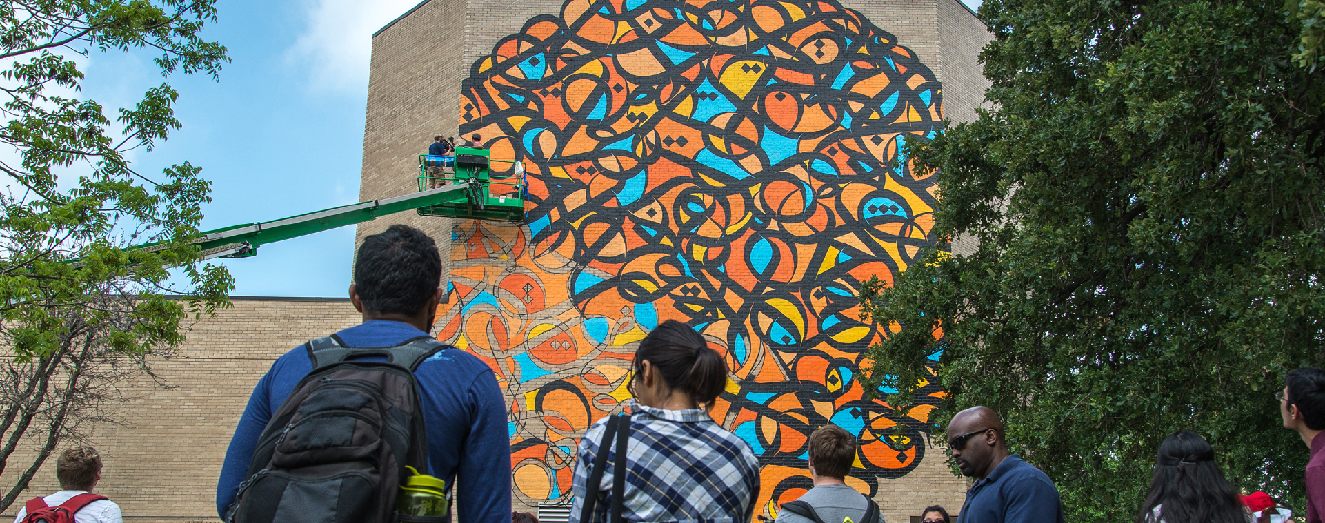 Two people in a lift paint a large orange, blue and black calligraphic mural on the side of a several-story high brown brick building while onlookers watch from below.