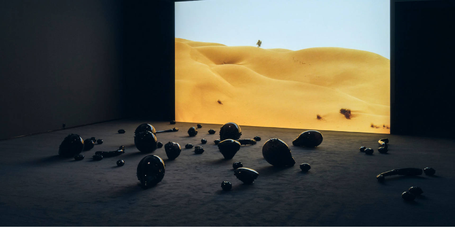 A dark room with rounded, shiny, black sculptures arranged on the floor in front of a projection screen showing a landscape of large yellow sand dunes and blue sky.