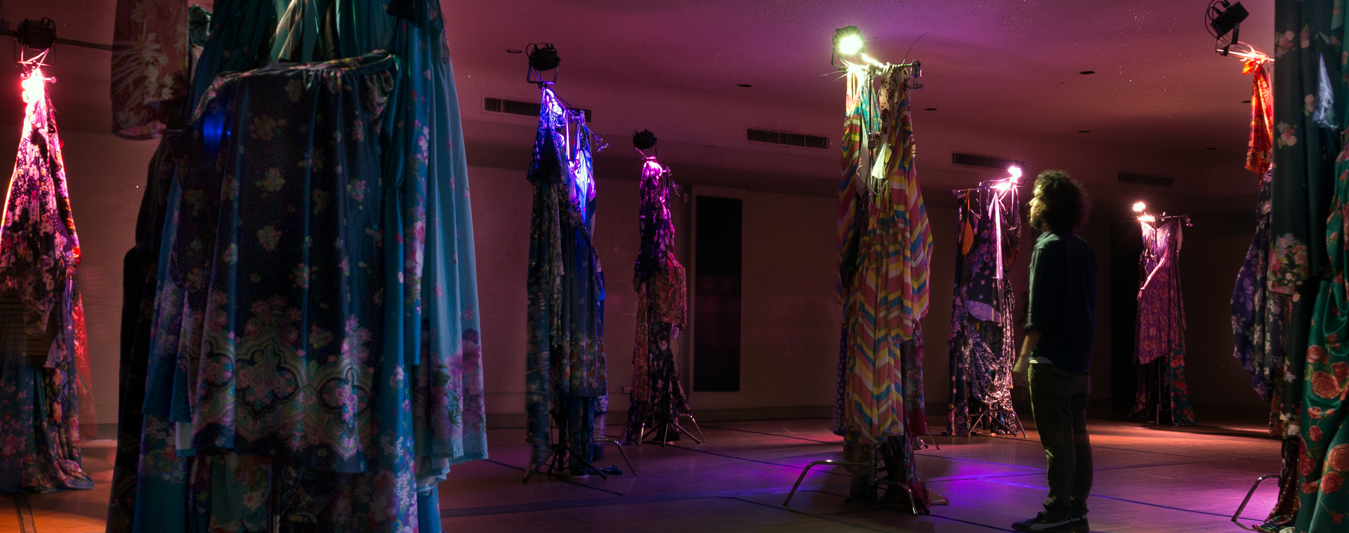 An art installation in a dimly lit room with patterned fabrics adorned on tall poles, each topped with a colored light; a person turned away from the camera stands in front of one.