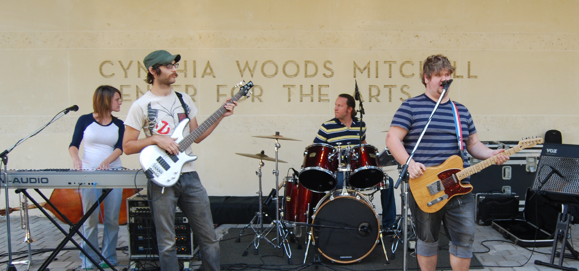 A rock band plays in front of the Cynthia Moods Mitchell Center for the Arts building signage, two people play guitar, one plays they keyboard and the other plays the drums with sound equipment behind them.
