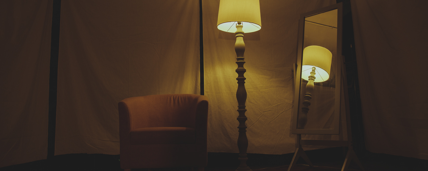 A floor lamp is reflected in a mirror to its right which lights an armchair situated in the middle of a cloth partitioned room with wood plank flooring.