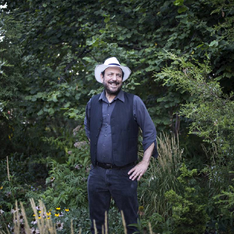 A smiling white-skinned man wearing a white hat and dark outfit standing amid lush greenery with a wall of trees behind him.