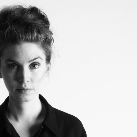 A black and white headshot of a white-skinned woman against a light backdrop with dark hair styled on top of her head, one eyebrow slightly raised, wearing a dark collared shirt.