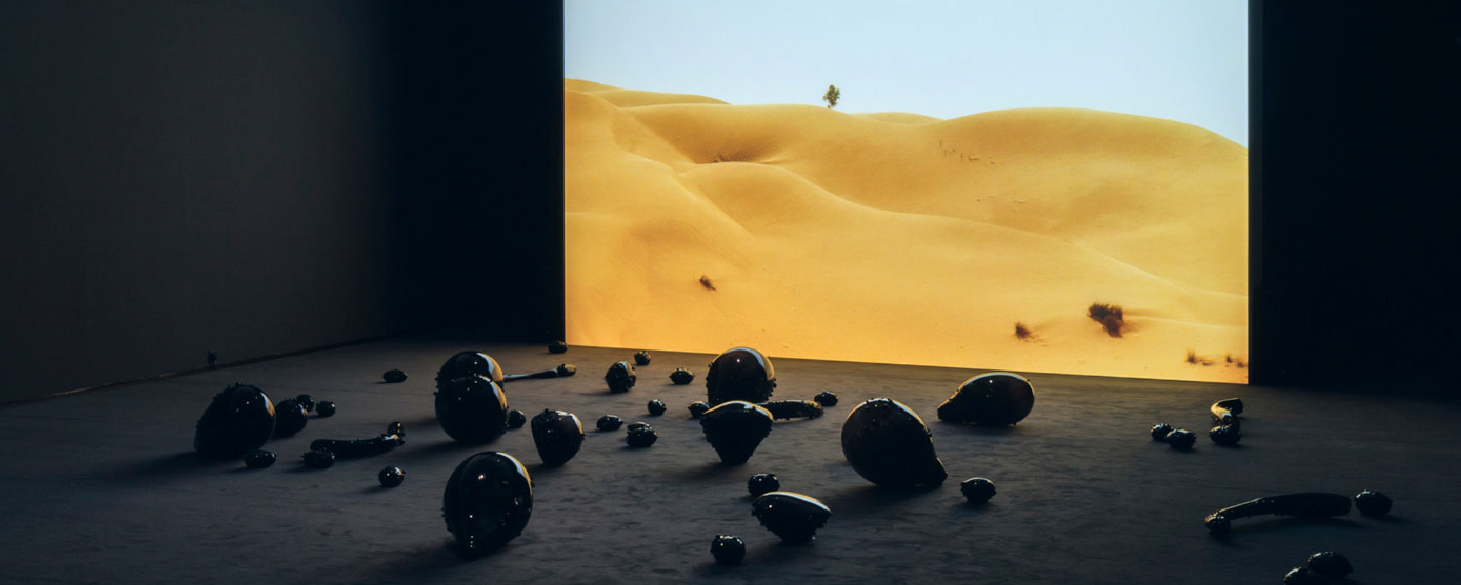 A dark room with rounded, shiny, black sculptures arranged on the floor in front of a projection screen showing a landscape of large yellow sand dunes and blue sky.