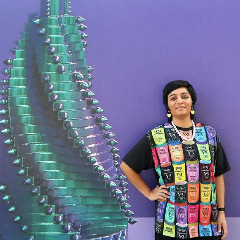 A smiling, brown-skinned woman with short black hair wearing a multicolored T-shirt stands in front of a purple wall beside an oversized spiraling green tower.