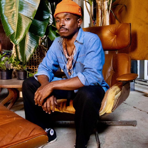 A black-skinned man with a mustache, sitting, leaning forward in a tan leather chair, wearing a beanie and blue collared shirt in an interior space with large plant leaves in the background.