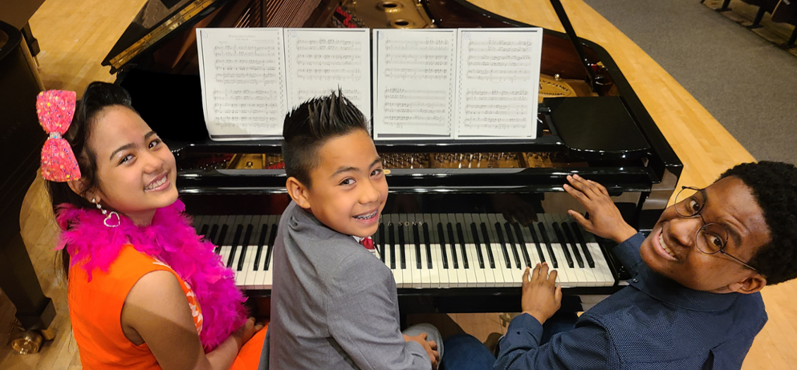 three young students sitting at a piano, turned around and smiling at the camera