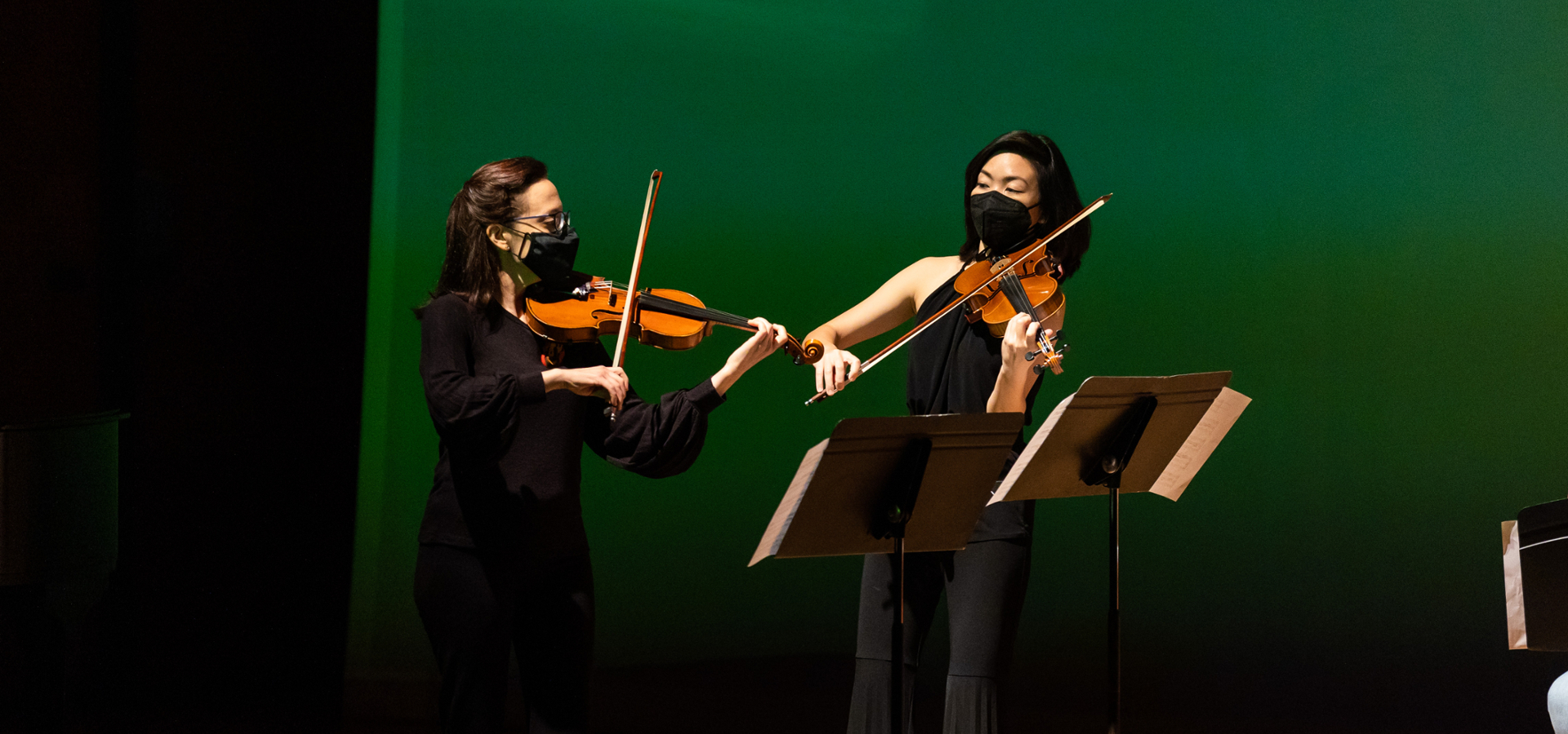 Dr. Kirsten Yon and Dr. Mann-Wen Lo playing violin together in front of a turquoise background.