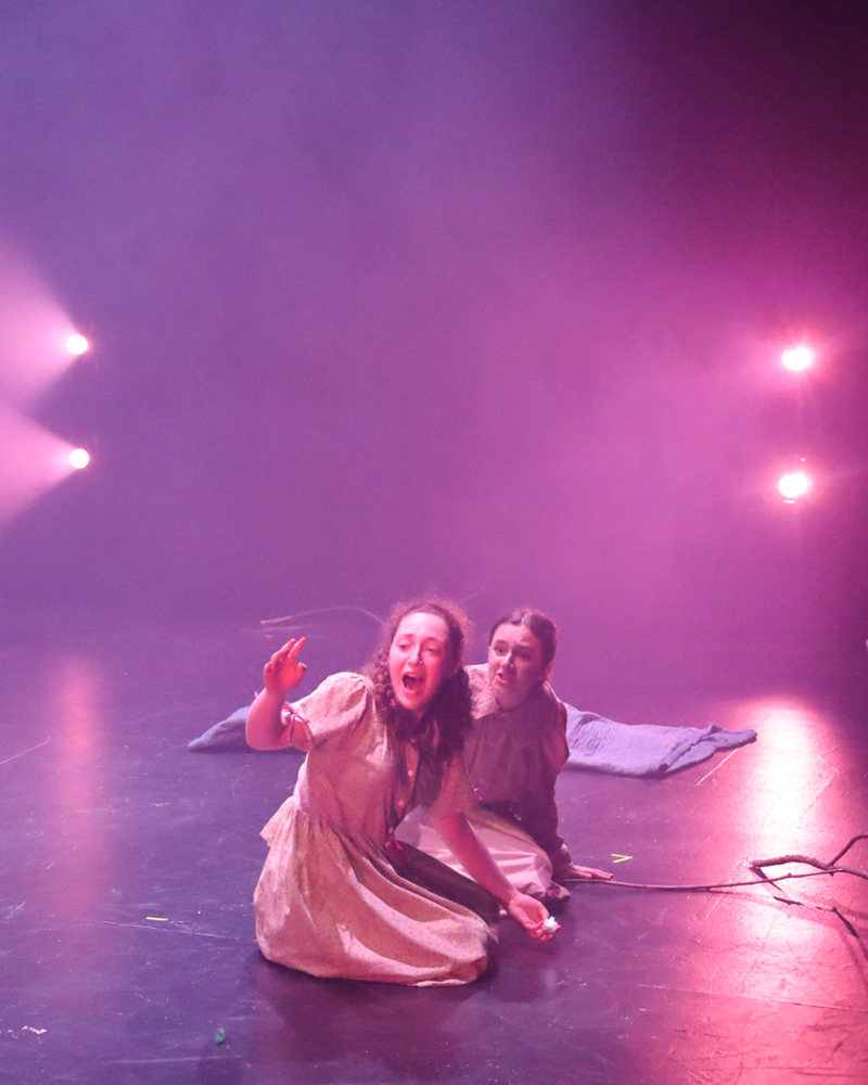 Two actors kneeling on the ground and yelling at someone off-stage, surrounded by lights of pink and purple