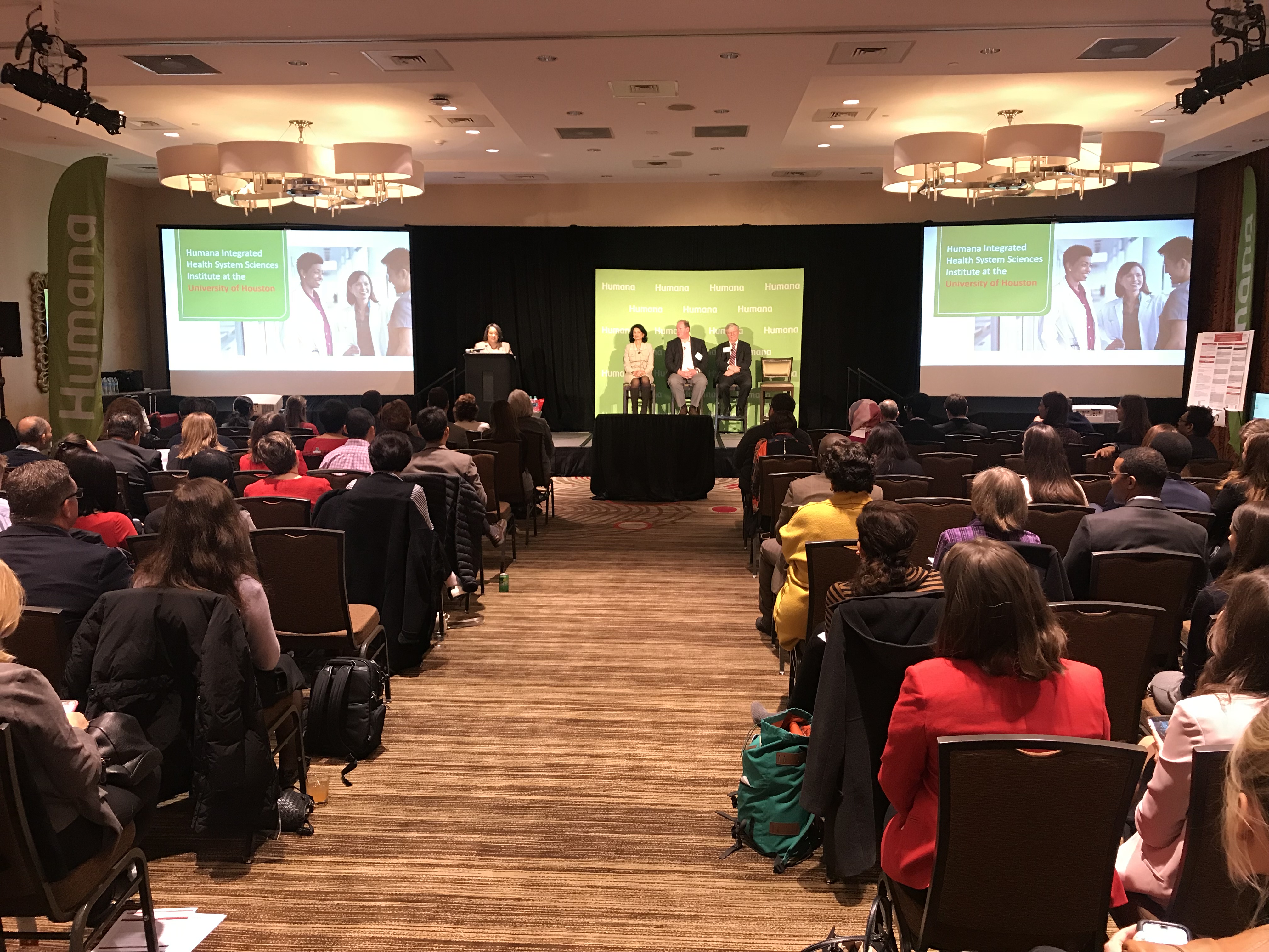 wide shot of room, backs of heads. Panel on stage at front.