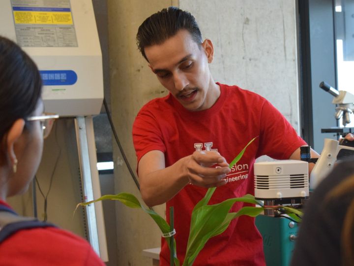 Person in a red University of Houston shirt conducting a scientific demonstration with plants and laboratory equipment.