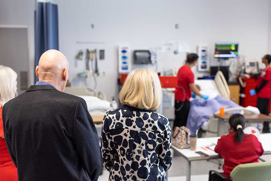 Photo of the backs of the two women and one man. They look towards a groupd of nursing students practicing on a mannekin.