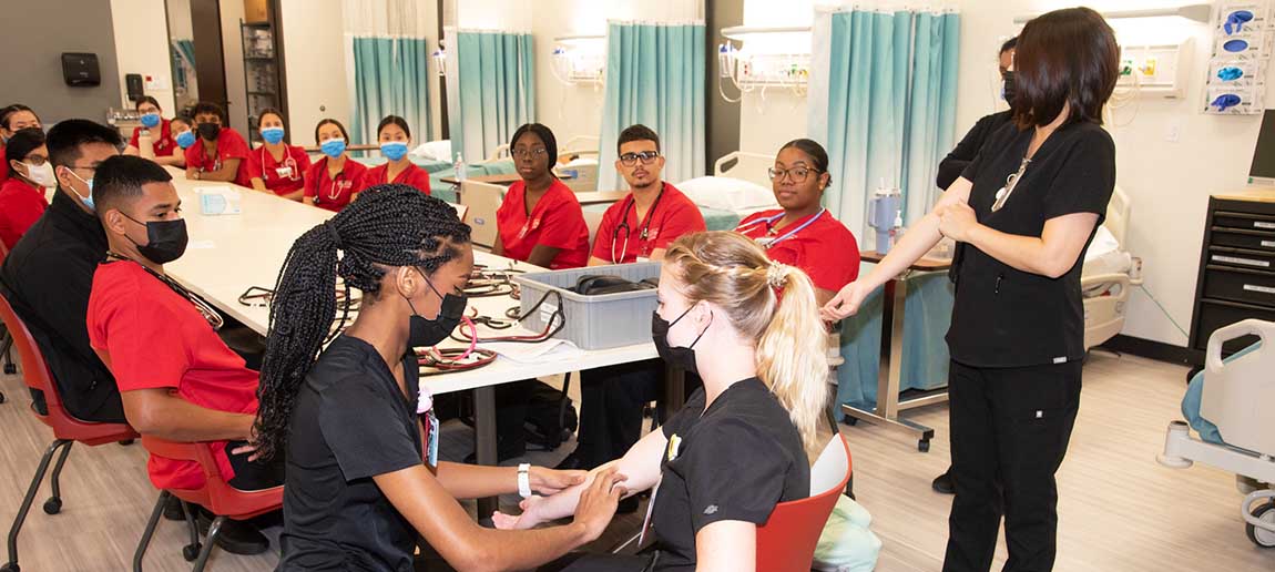 A group of nursing student watch as a instructor explains a demostration with two students in a lab.