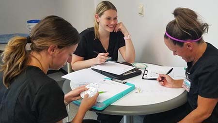 Three female nursing students sit at a table together writing on notepads.