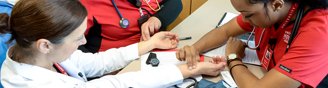 A nursing student looks at her watch while she measures the pulse of her instructor.
