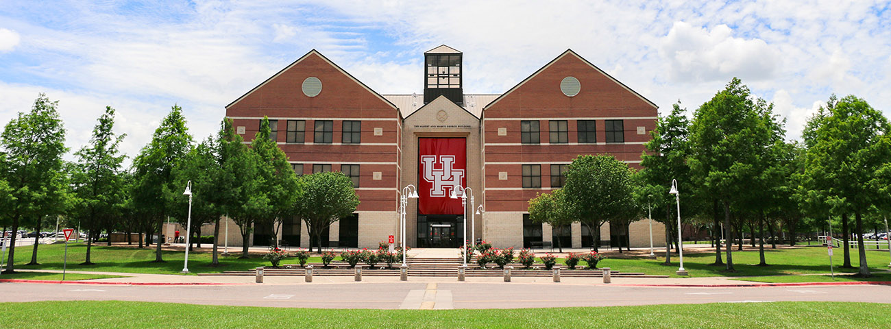 A wide shot of a large three-story brick building with a large red UH banner on the front. Green trees surround the sides of the building.