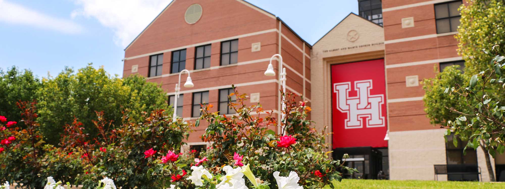 The front of a brown building with a red and white University of Houston banner with trees and bushes in front of it.