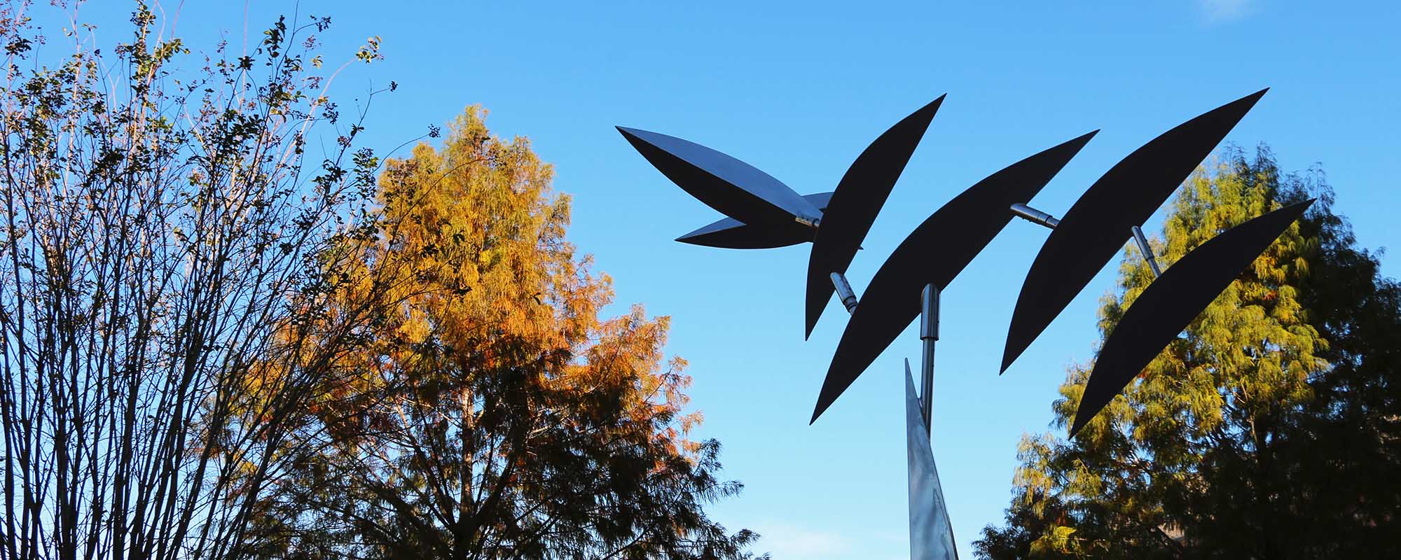 A metallic sculpture with leaf-like parts in the foreground and trees with red and green leaves in the background