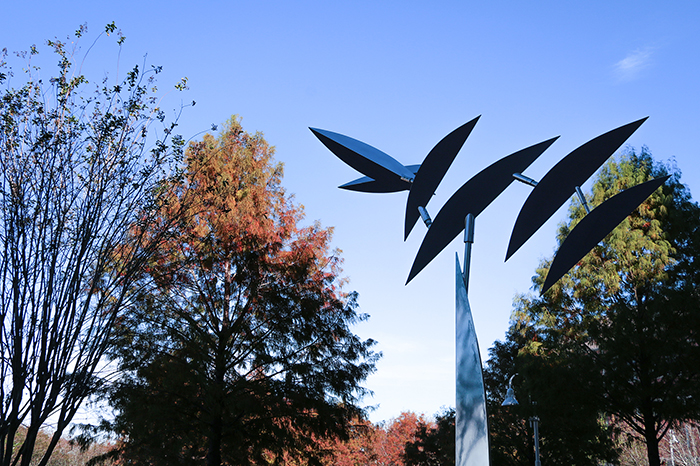 A metalic sculpture with leaf-like parts in the foreground and trees with red and green leaves in the background