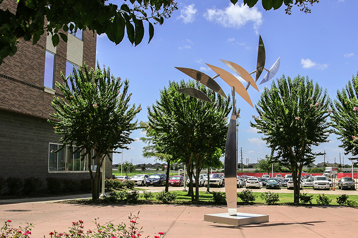 A metalic sculpture with leaf-like parts in the middle of a redish brown patio. Green trees and a brown multistoried building are in the background.